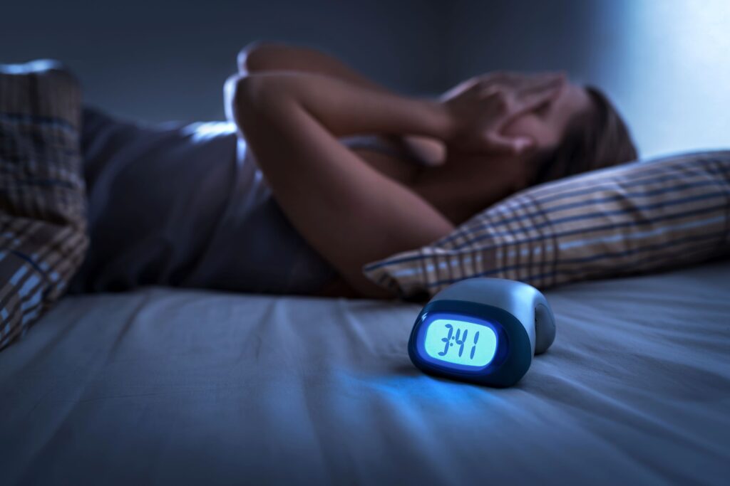 Woman in bed next to alarm clock with hands over face frustrated